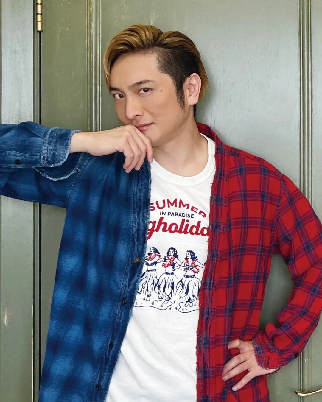 金髪の男性が、青と赤のチェック柄シャツを羽織り、フラダンスのイラストが描かれたTシャツを着てポーズ