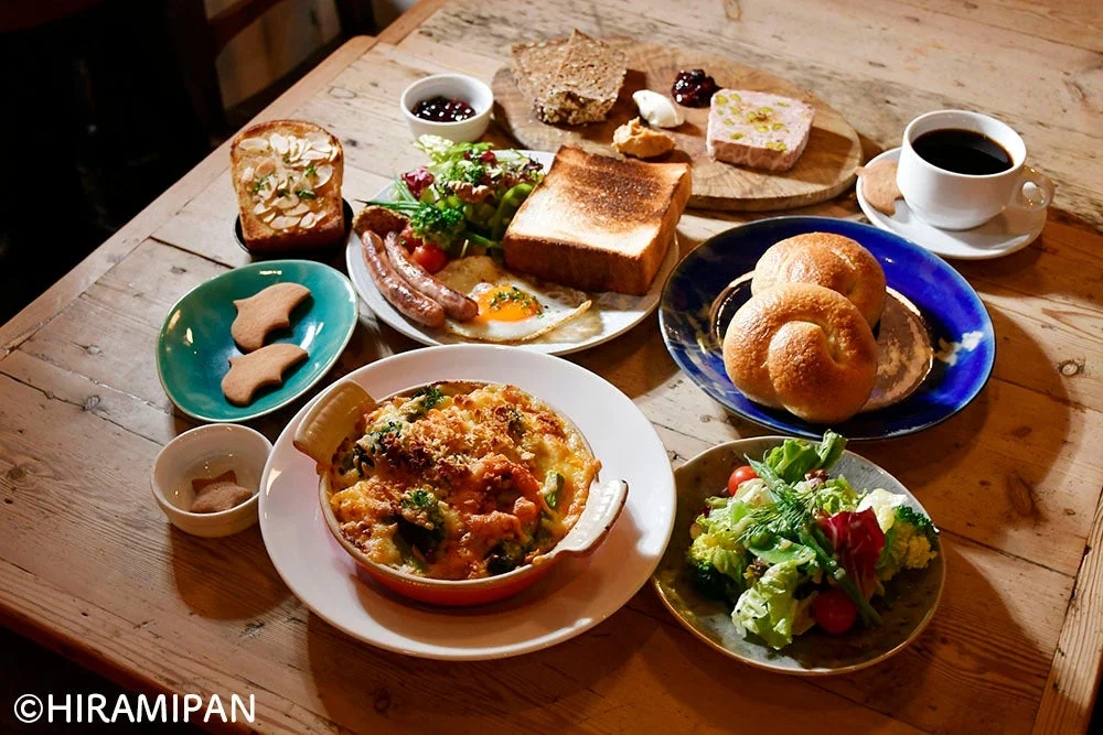 木製のテーブルに、トースト、ソーセージ、目玉焼き、サラダ、グラタン、パン、コーヒー、パテなどが並んだ豪華な朝食またはブランチの風景です。