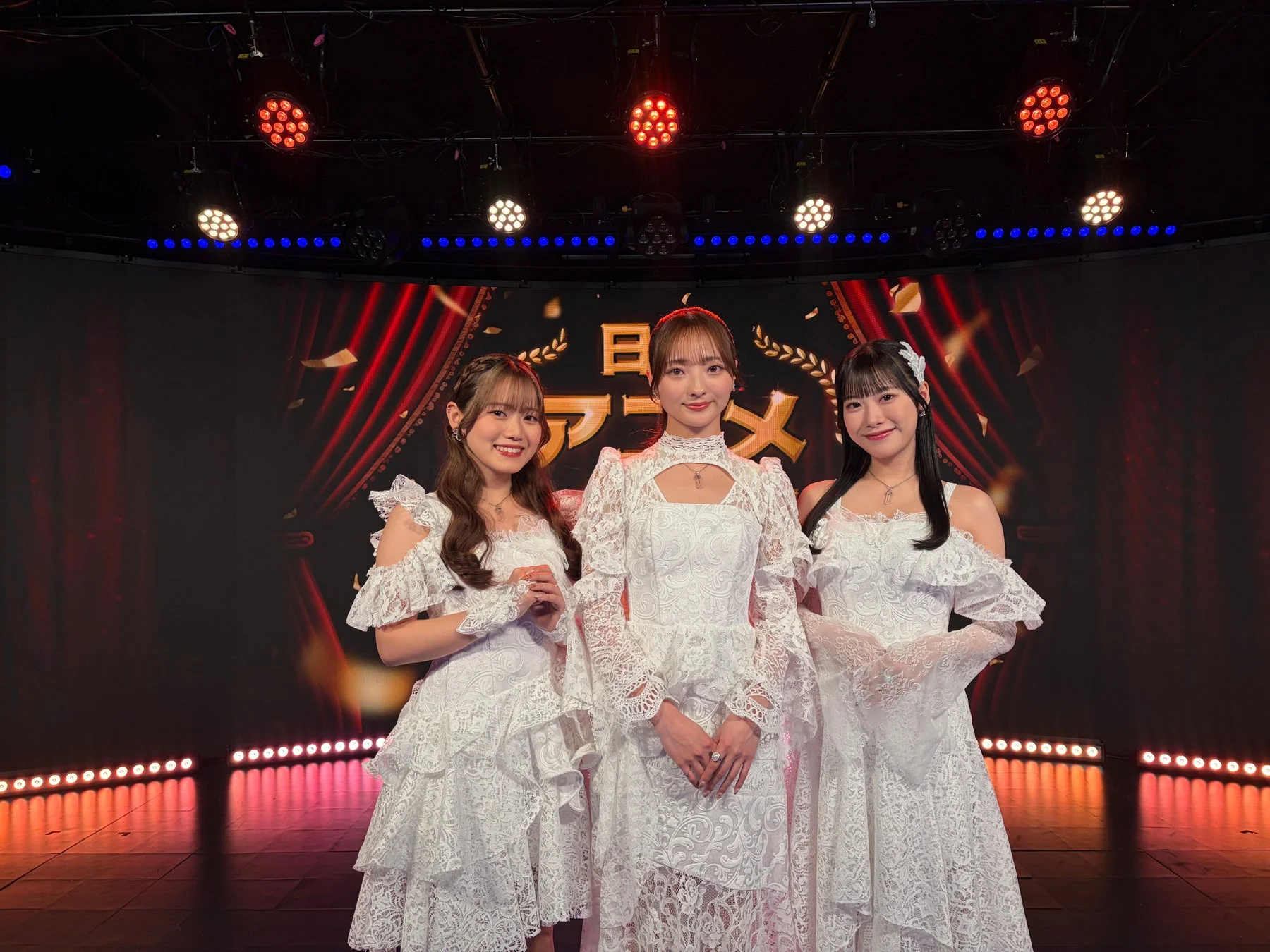 白いレースのドレスをまとった3人の女性が、ステージ上で笑顔を見せている