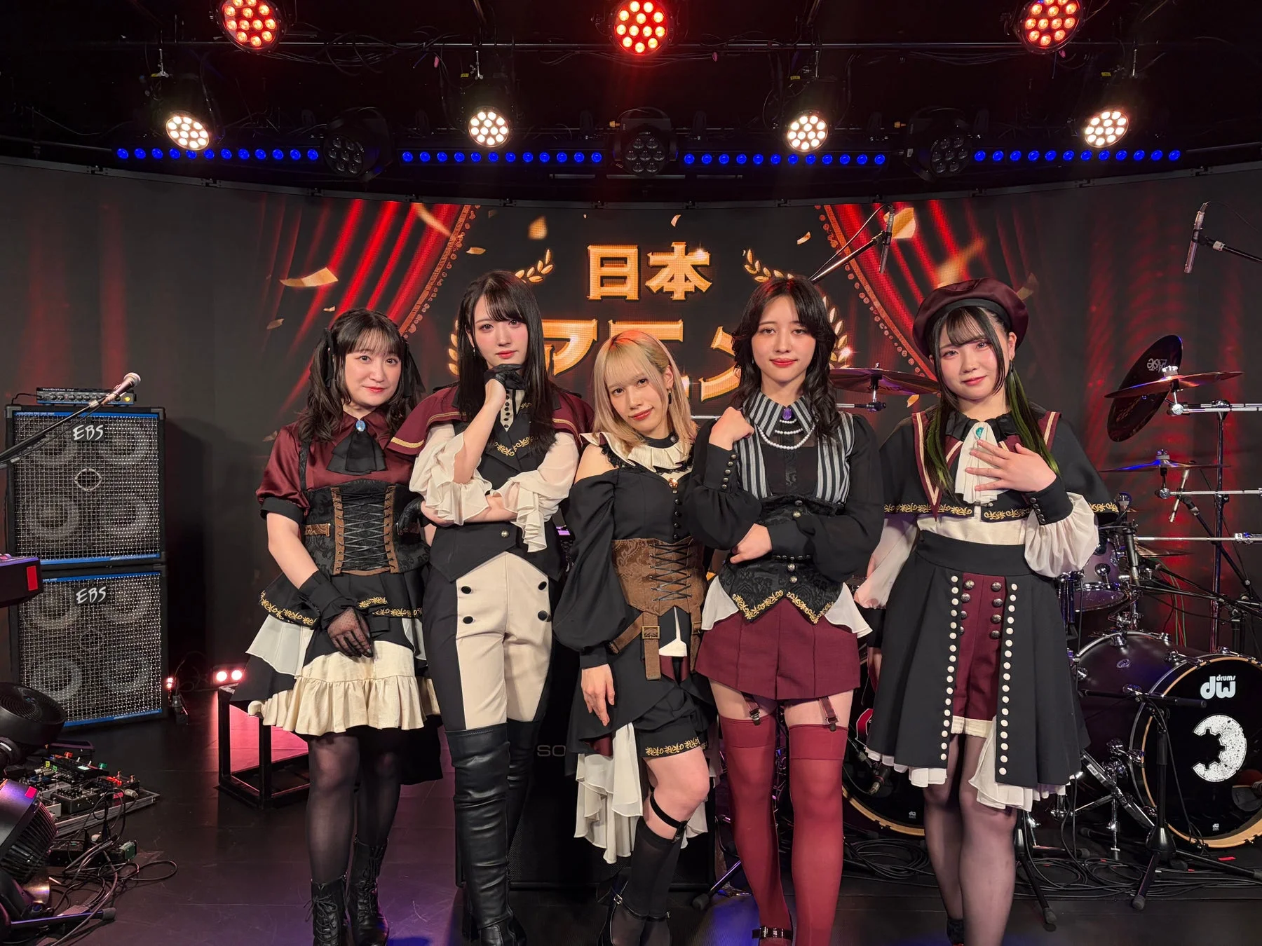 個性的なステージ衣装をまとった5人の女性が、明るい照明のライブステージでポーズをとっている