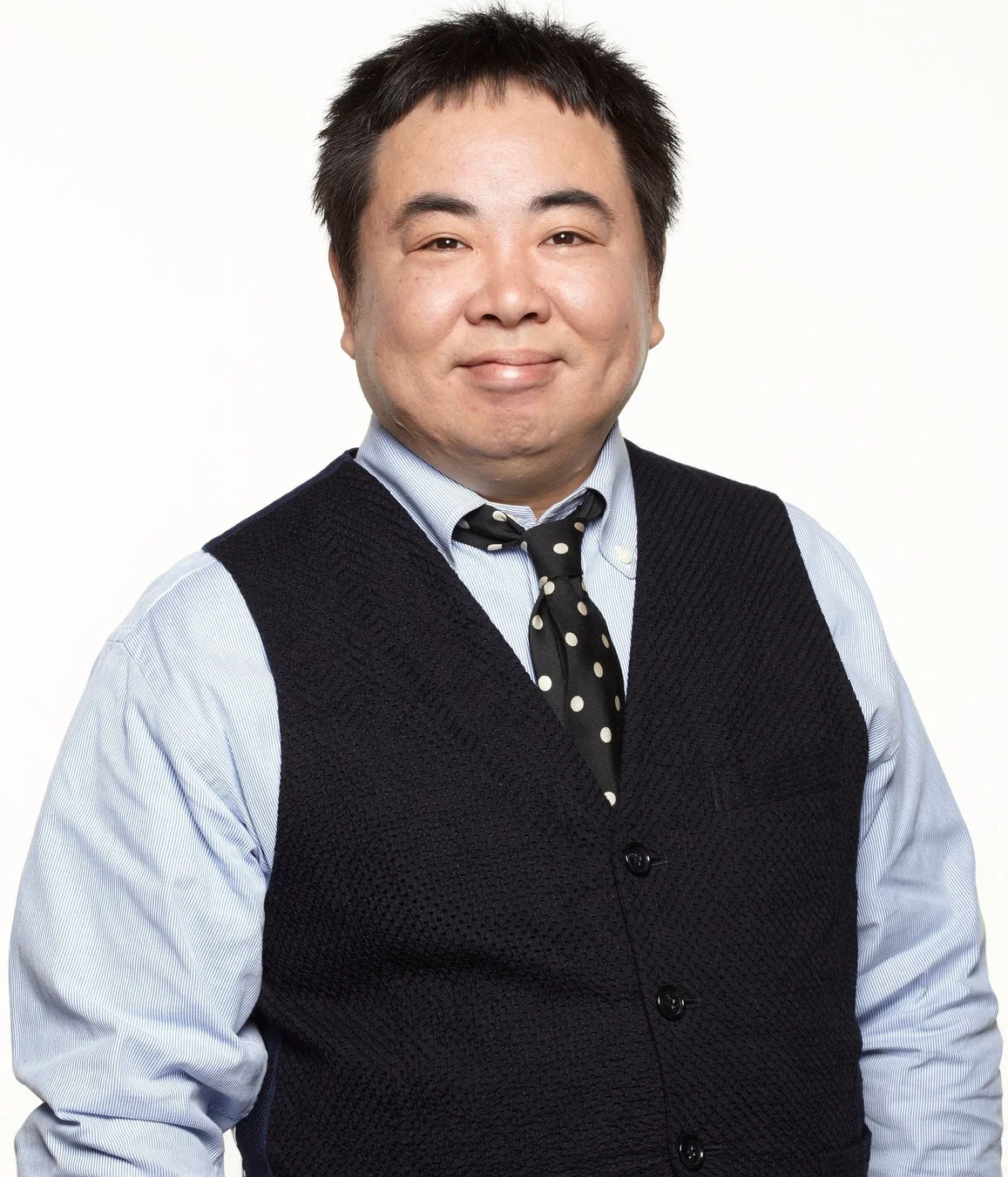 水色のストライプシャツと黒いベスト、水玉模様のネクタイを着用したアジア人男性がカメラに向かって笑顔