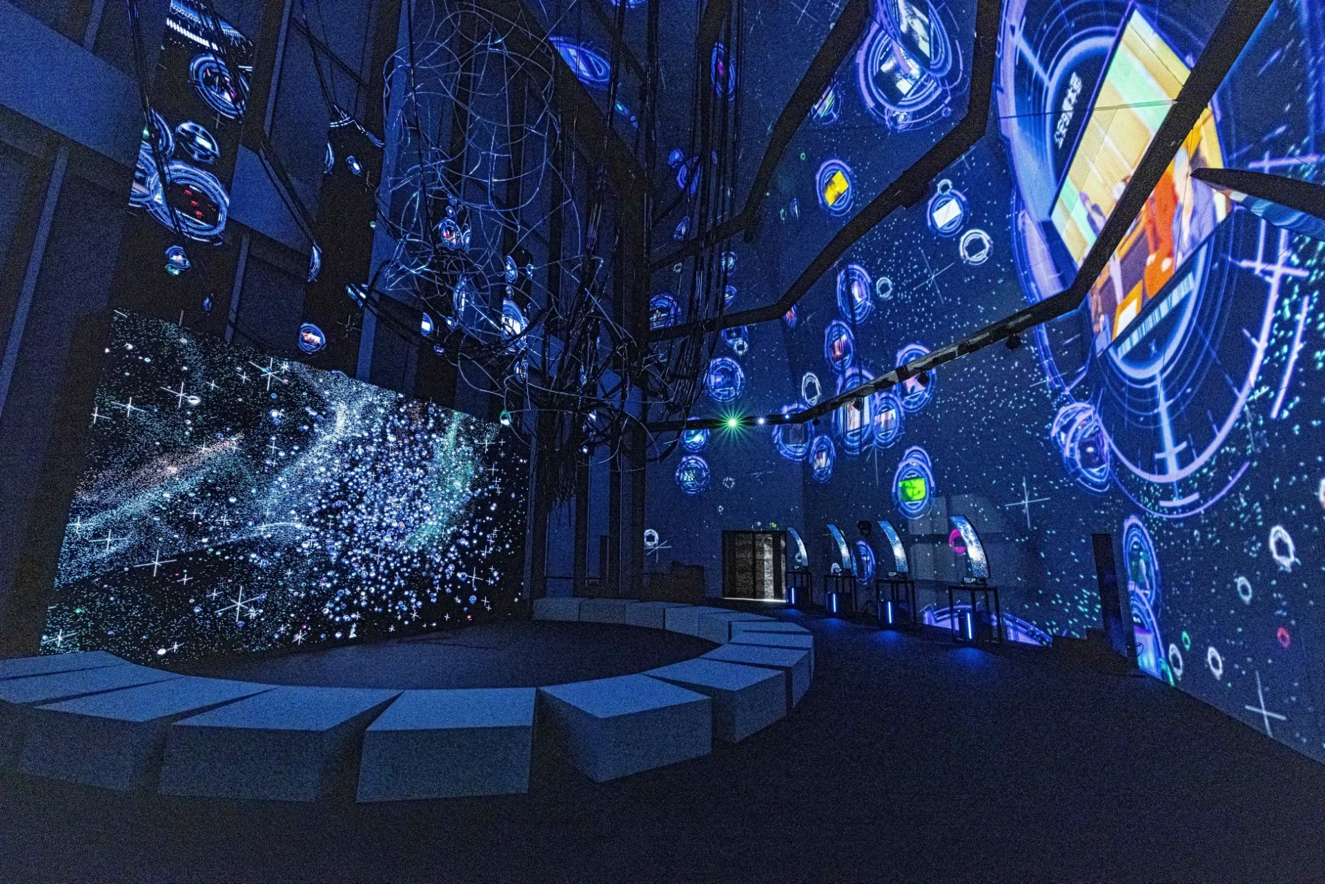 青い光が印象的な未来的な展示空間。壁面には星空やデジタルパターンが投影され、中央には湾曲した座席がある。来場者が没入感のある体験を楽しめる科学館やアート展示の様子。
