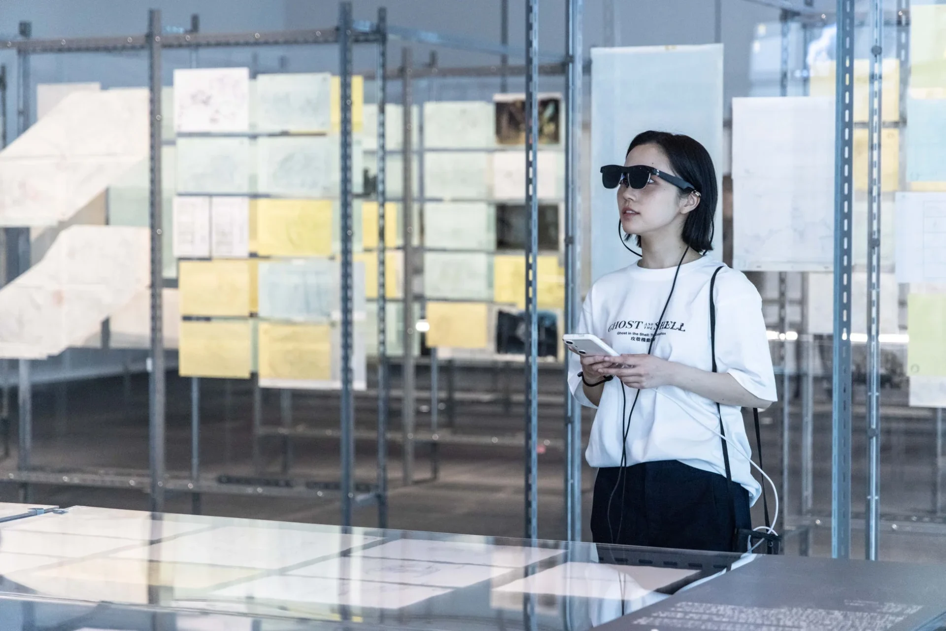 スマートグラスを装着した女性が、多数の資料が展示されたギャラリーでスマートフォンを手に鑑賞しています。彼女のTシャツには「攻殻機動隊」の文字が見えます。