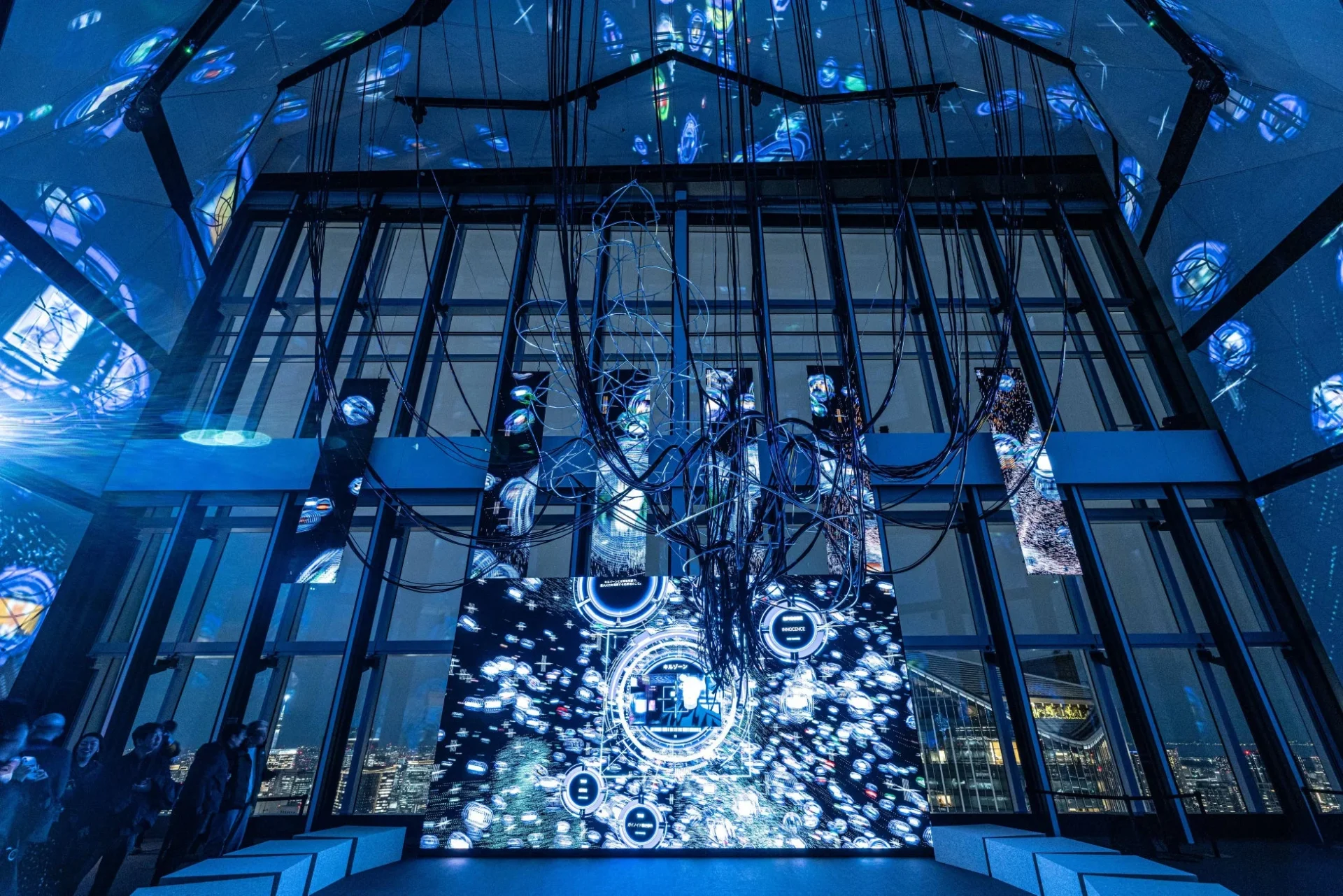 青い光とデジタルアートが特徴的な未来的な空間で、天井からは無数のケーブルが垂れ下がり、大きなスクリーンには抽象的なパターンと情報が表示されている。窓からは夜景が広がり、人々がこの没入型インスタレーションを鑑賞している様子。
