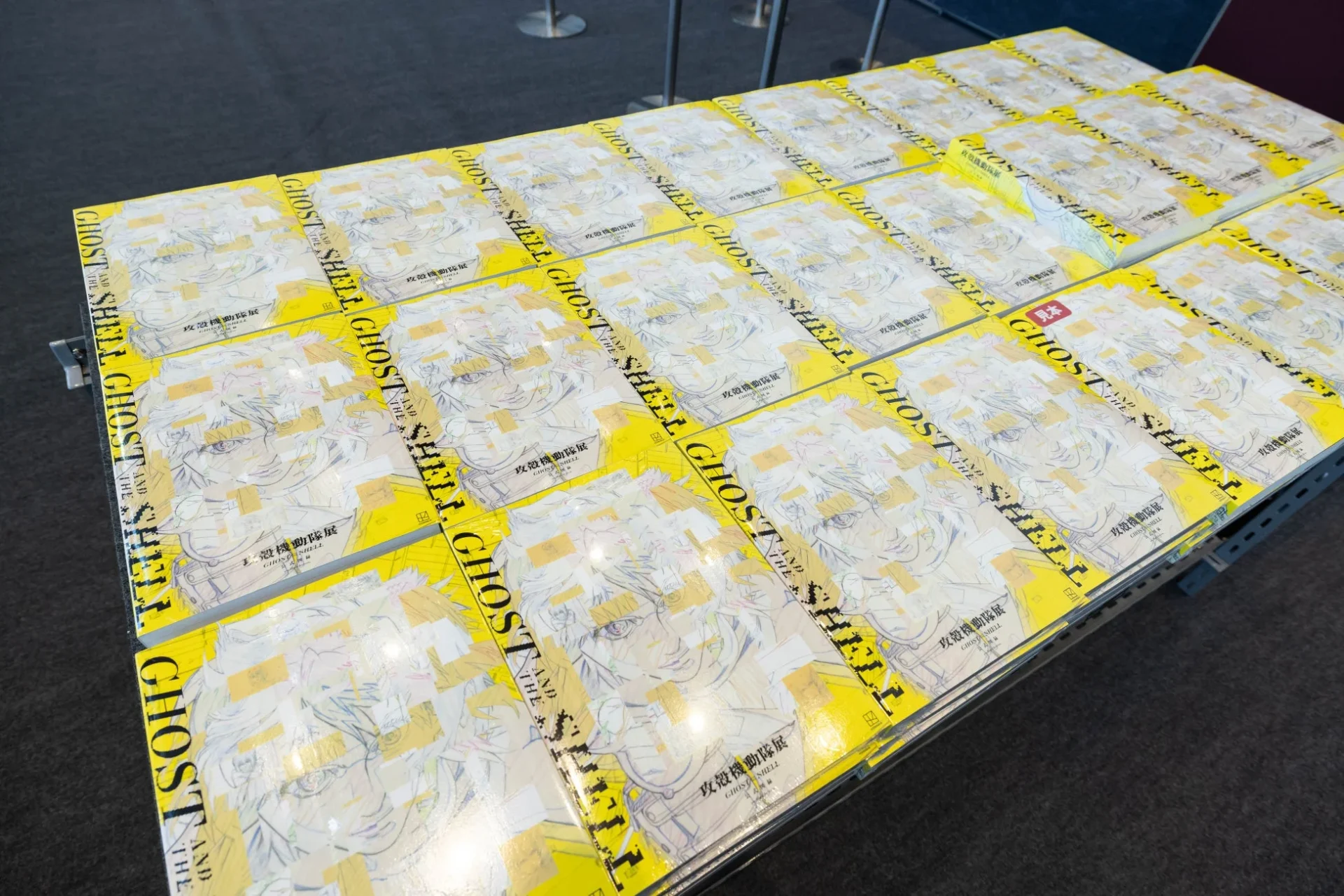「攻殻機動隊展」の会場で、黄色い表紙の「GHOST IN THE SHELL」関連書籍が多数並べられています。表紙には主要キャラクターのイラストが描かれています。