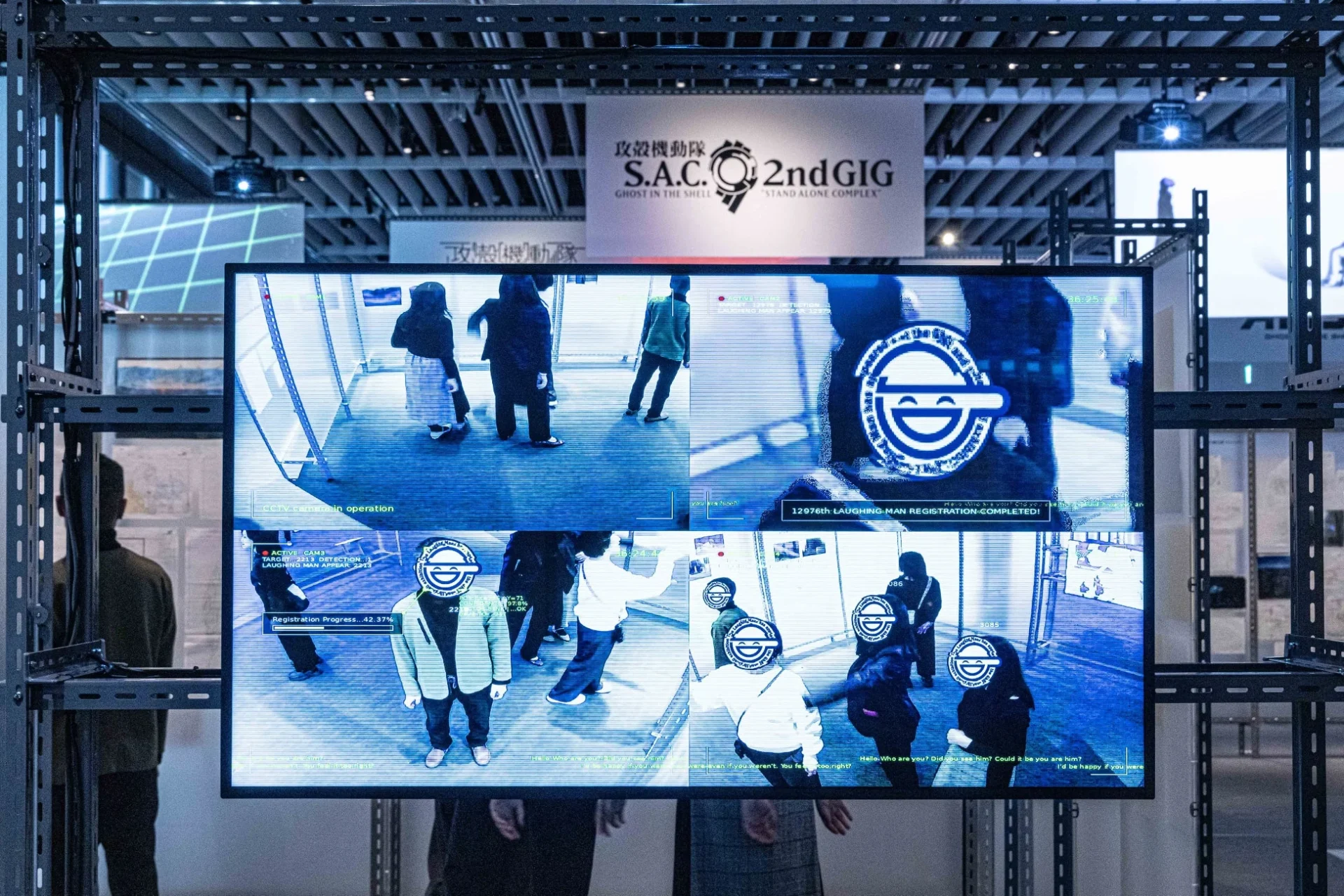 「攻殻機動隊 S.A.C. 2nd GIG」の展示風景で、監視カメラのようなスクリーンに「笑い男」のロゴが映し出され、来場者の顔に重ねられている様子が描かれています。