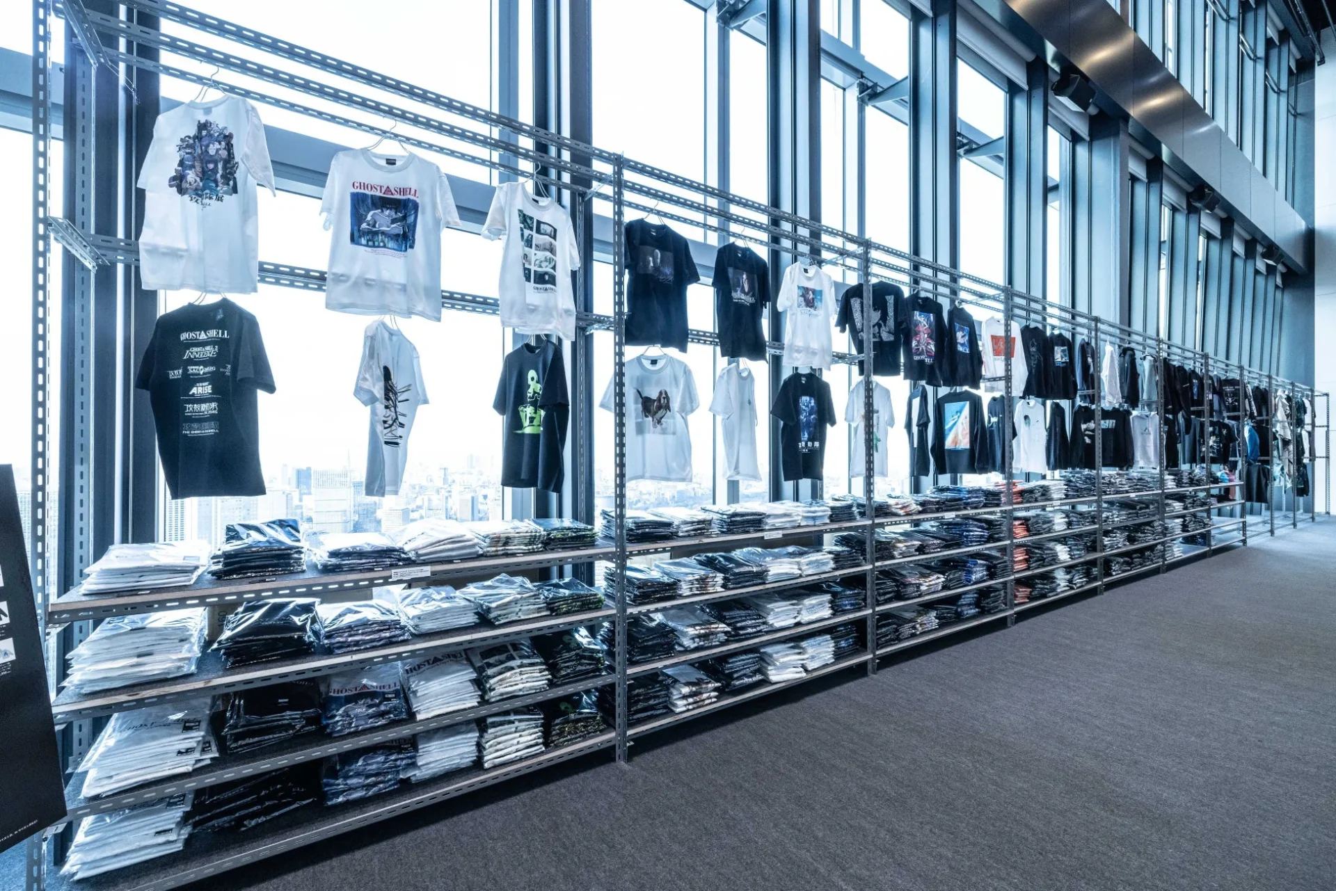 高層ビルの窓際に設置された棚に、人気アニメ「攻殻機動隊」シリーズのTシャツが多数展示・販売されている様子。白と黒を基調としたTシャツがハンガーに吊るされ、下の棚には畳まれた商品が並び、都会の景色を背景にしたポップアップストアのようだ。