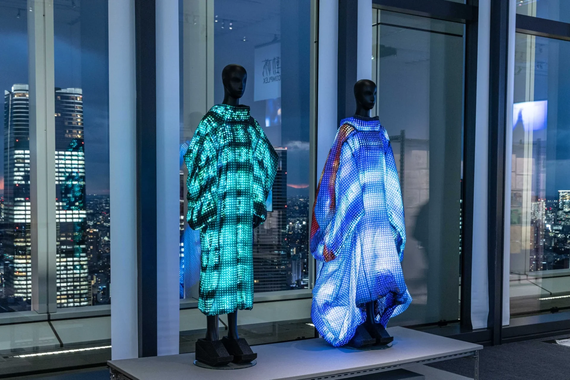 夜景が見える高層階の窓際に、LEDで光る未来的な衣装をまとった2体のマネキンが展示されています。緑と青の光が特徴的なアート作品です。