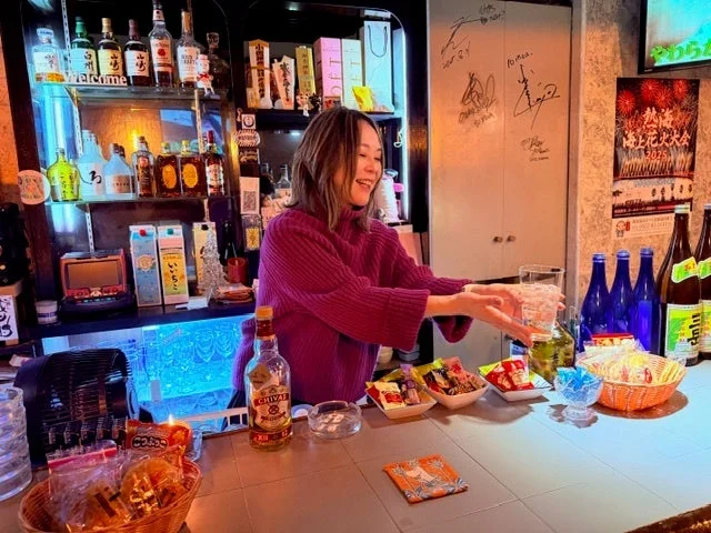 バーカウンターで笑顔の女性が接客しており、カウンターには様々なお酒のボトルやスナックが並ぶ