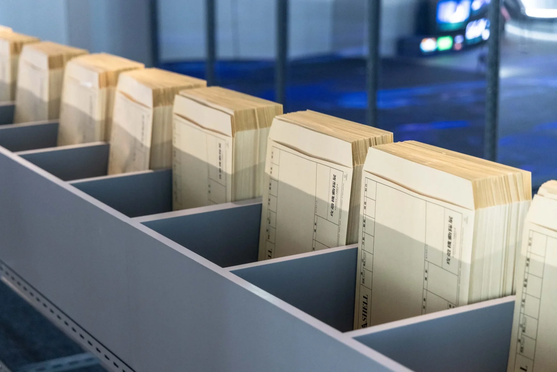 灰色に仕切られた棚に、古い日本語の書類が何束も整然と並べられています。書類には「政設機動隊」などの文字が見え、歴史的な資料が展示されている様子が伺えます。