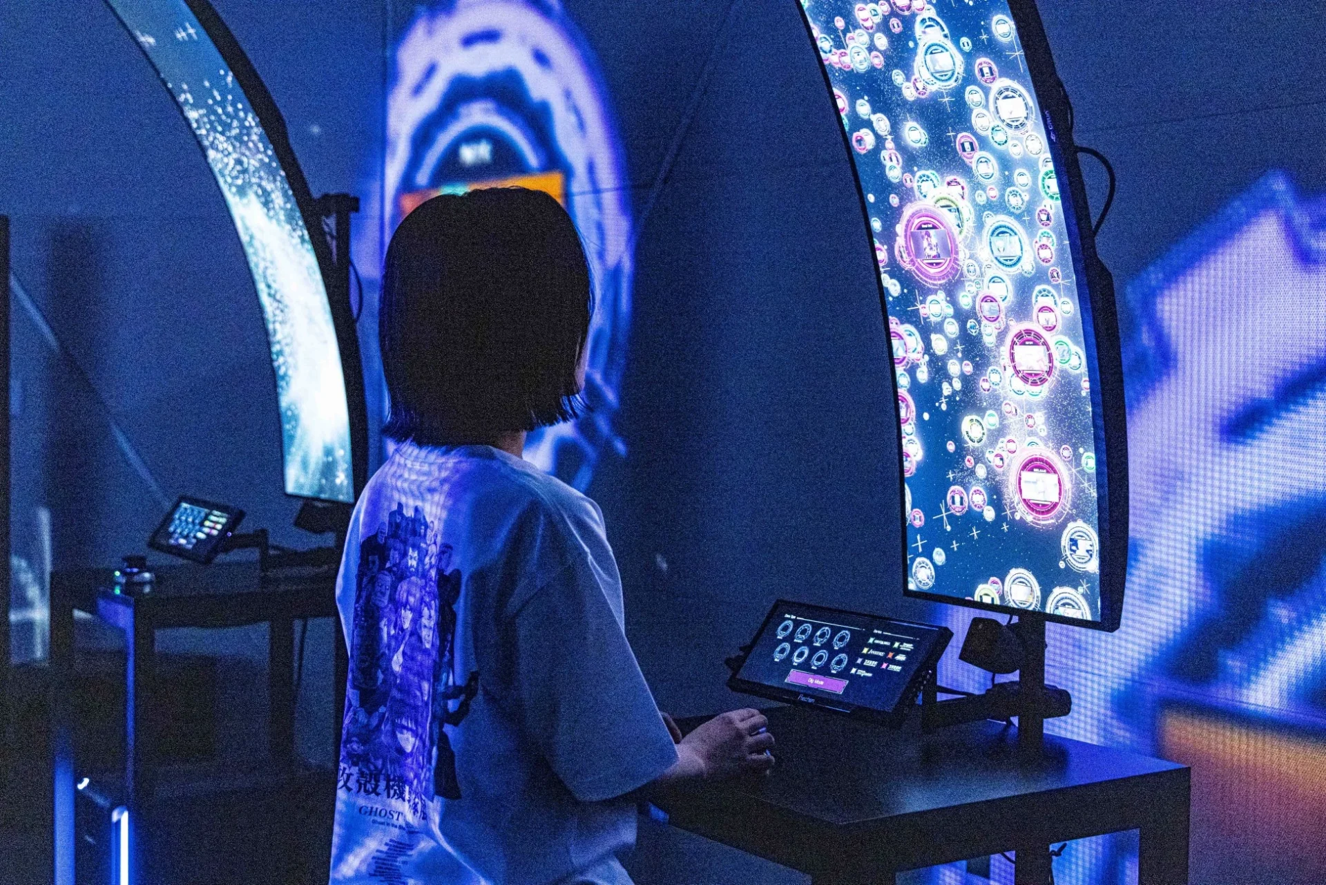 青と紫の光に包まれた空間で、一人の人物が未来的なデジタル展示を体験している様子。背後から見た人物は「攻殻機動隊」と書かれたTシャツを着用し、目の前の大型カーブモニターと操作用タブレットに集中している。没入感のあるサイバーな雰囲気が特徴的。